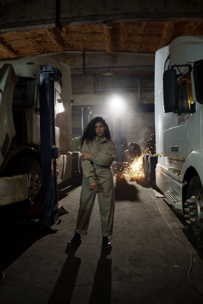 Confident female mechanic standing in a truck repair workshop with sparks flying in the background.